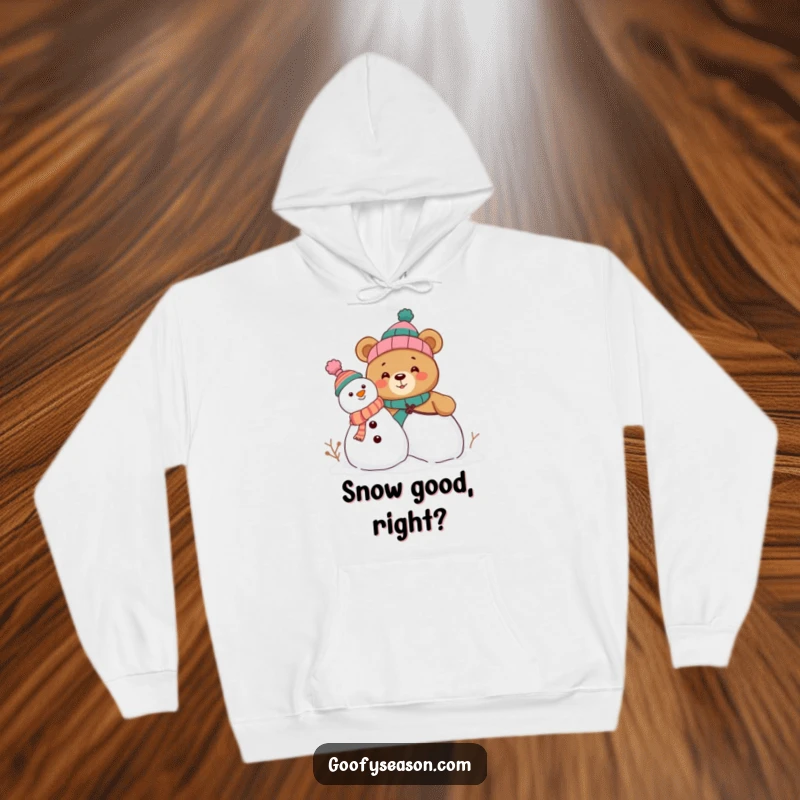Funny hoodie showcasing a curious bear character constructing a snowman with a pile of mismatched, funny hats, radiating winter cheer.